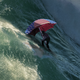 Detail of illustration of person holding large umbrella, with alternating stars and stripes of the U.S. flag, climbing the steep face of an enormous ocean wave breaking overhead.