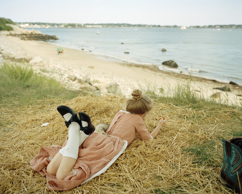 Photo of a young woman in pink dress lying on stomach, with legs crossed wearing long white socks and black shoes, in pile of straw looking out at the beach and water at Gloucester, MA