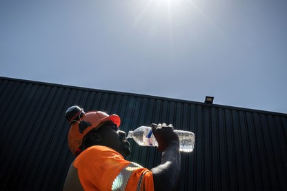 A construction worker in an orange hat and shirt drinks from a liter bottle of water under the glaring sun
