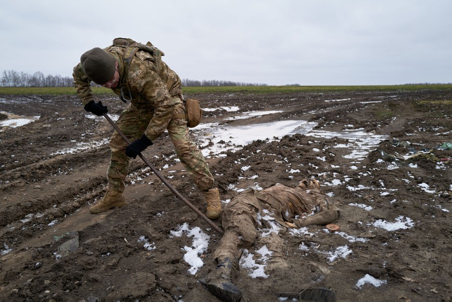 A Ukrainian soldier uses a steel bar to pry the body of a Russian soldier from frozen mud.