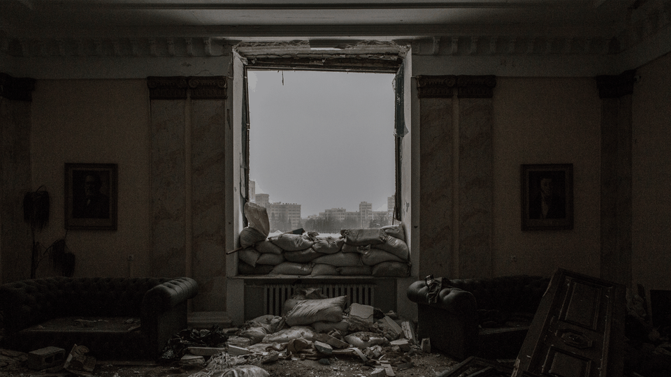 A photograph of a bombed-out room with sandbags piled behind a glassless window