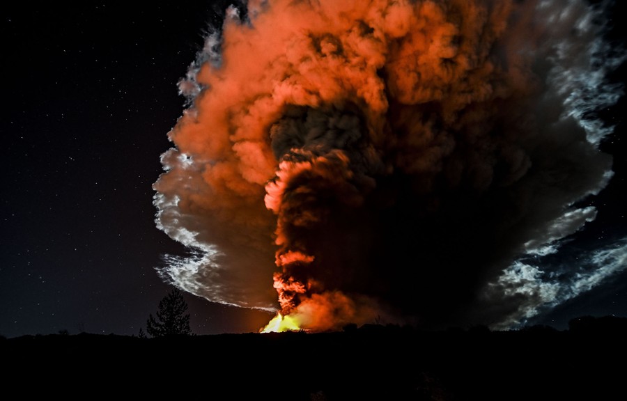A night view of a volcano sending a huge cloud of ash into the sky, lit from below by lava glow.
