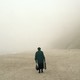 Color photograph of the back of a person in black clothing, carrying bags and standing on a beach