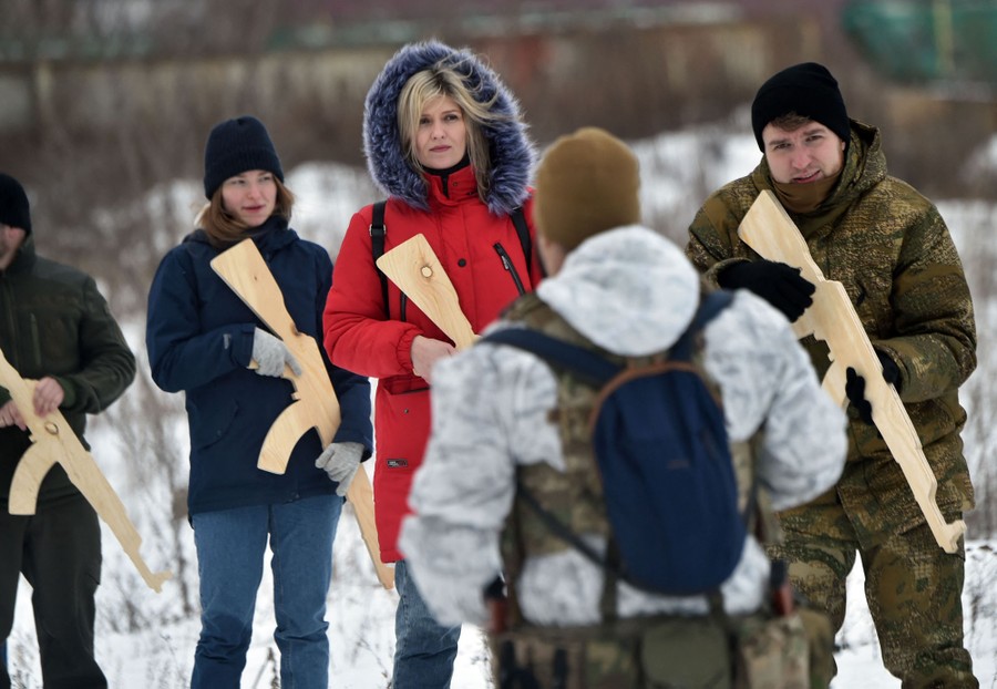 Civilians hold wooden replicas of rifles while being instructed, standing in a field.