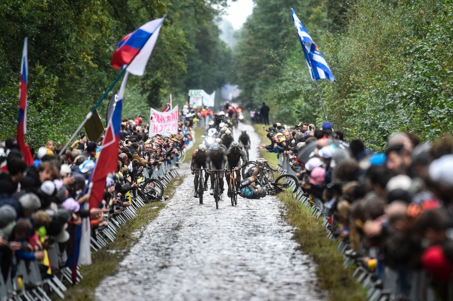 A view down a long cobblestone road, lined with racing fans, with a small group of cyclists riding in the middle and one on the ground after falling.