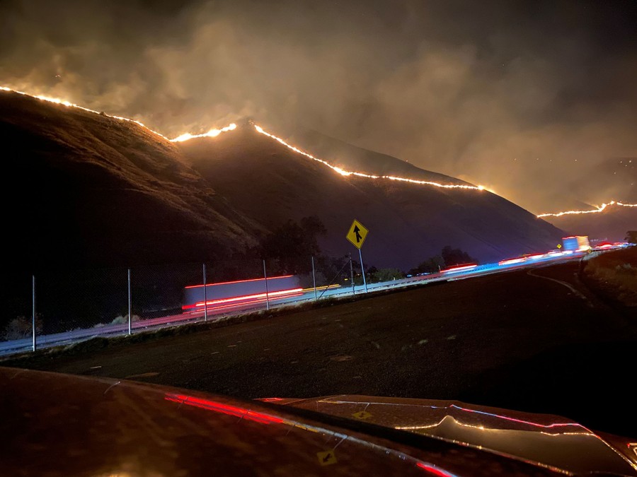 A wildfire burns in straight lines across several hillsides near a busy road.