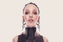 A color portrait of the singer FKA Twigs, seen head-on under bright light, wearing heavy eye makeup, big metallic earrings, and a sleeveless high-neck leather top.