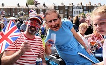 A man wearing Union Jack face paint and a woman wearing a King Charles face mask celebrate the coronation in a field while a child looks on, deeply distraught