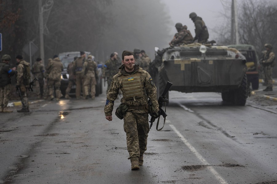 A soldier walks toward the photographer, while other soldiers stand in the background, on and near military vehicles.
