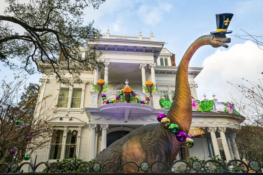 A large statue of a dinosaur wears a beaded necklace and top hat, standing in front of a decorated house.