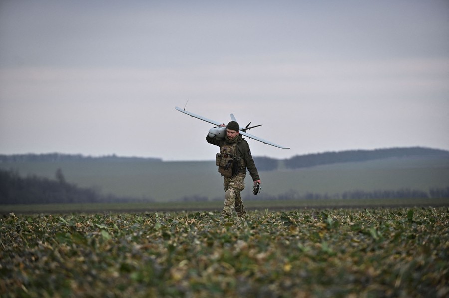A soldier walks through a farm field, carrying a drone over their shoulder.