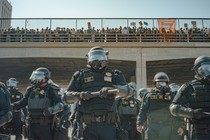 Police officers stand in a line in front of a bridge with people standing on it