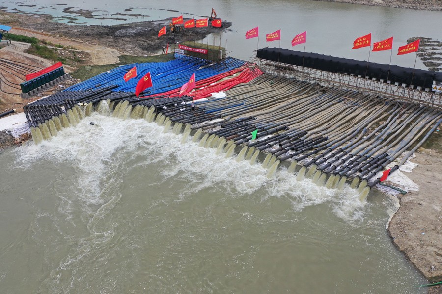 About a hundred large hoses lie side by side on an embankment between a river and a lake, each discharging water into the lake.