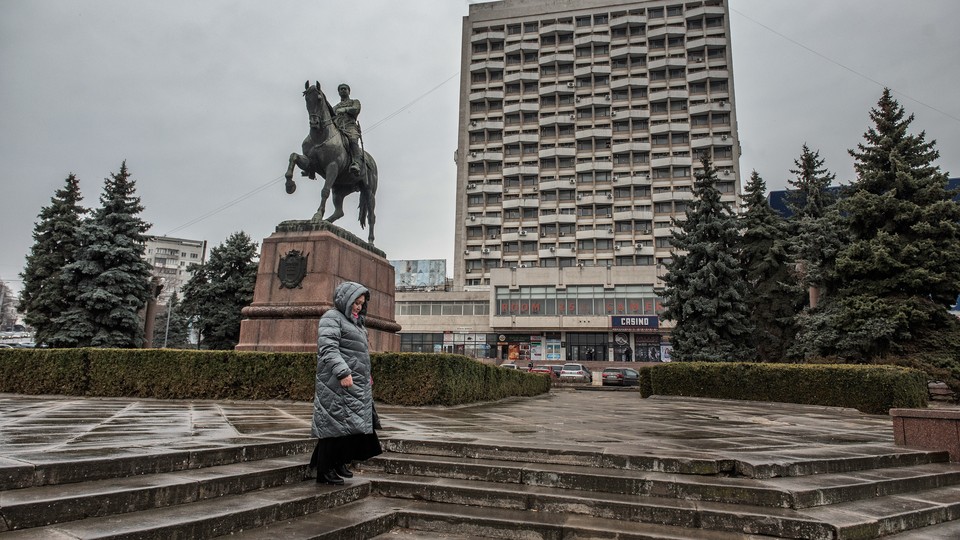Picture of a woman wearing winter rain gear and standing up in the stairs at a square with an equestrian statue and pine trees. In the background, there is a brutalist building with a sign that reads "Cosmos."
