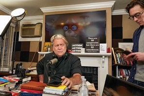 Photo of Steve Bannon sitting in cluttered home studio at microphone on desk covered with books near lighting equipment, in background a TV with still from the movie "Turning Red" over fireplace mantel with signs
