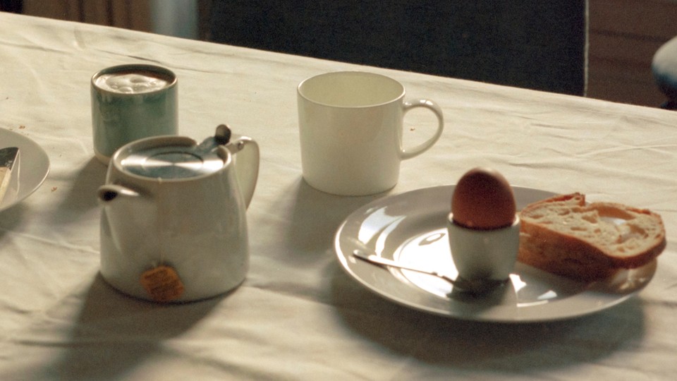 A table covered in a white table cloth, and on top, a tea kettle, two mugs, and a plate with an egg, a spoon, and a slice of bread