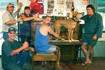 A group of men raise their beers while a dingo stands on a piano and howls.