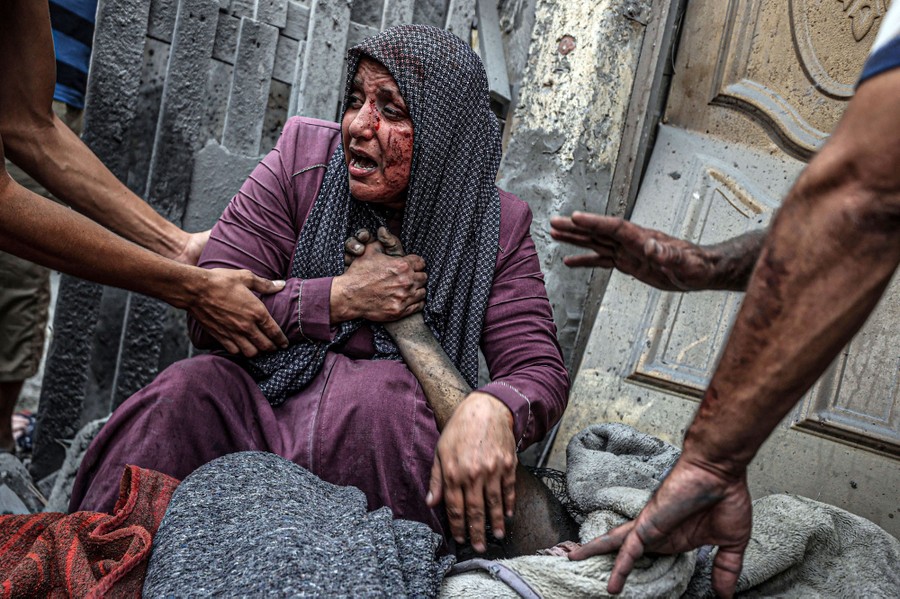 An injured woman cries beside a dead person, grasping their hand, as others lean in to help her.