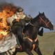 A rider on a horse, wearing flame-protective gear, rides with flames burning from their back, in a field.