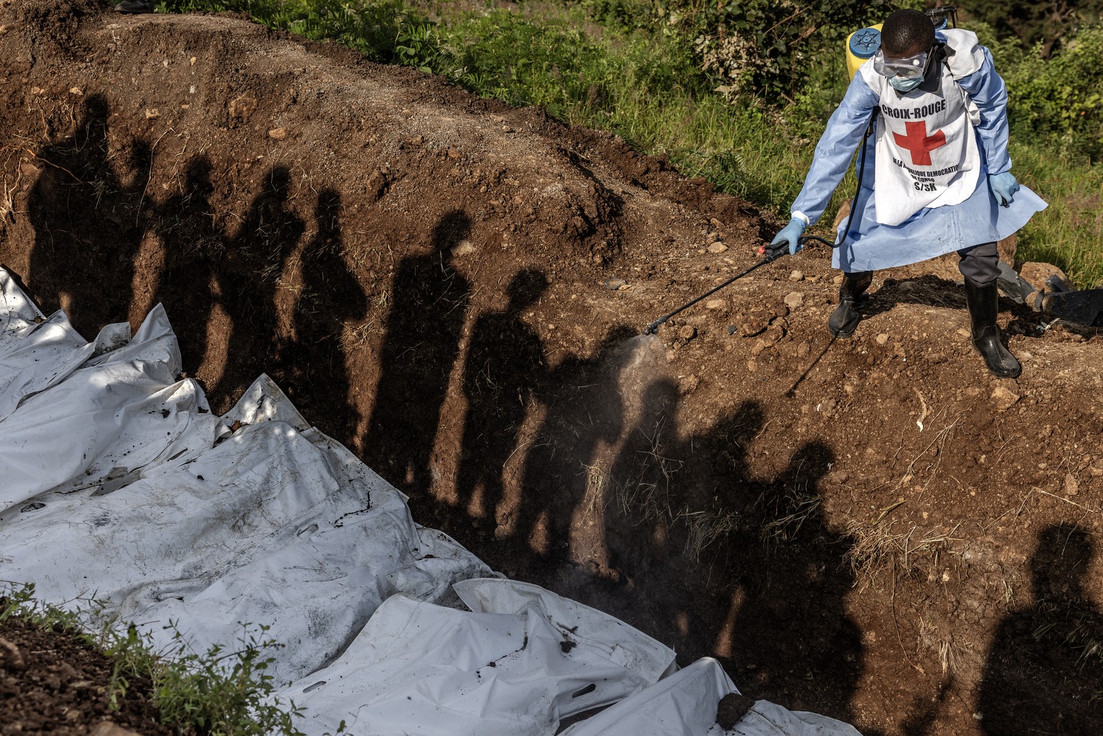 People's shadows fall along the wall of a mass grave, as a Red Cross worker sprays disinfectant on bodies in white bags.