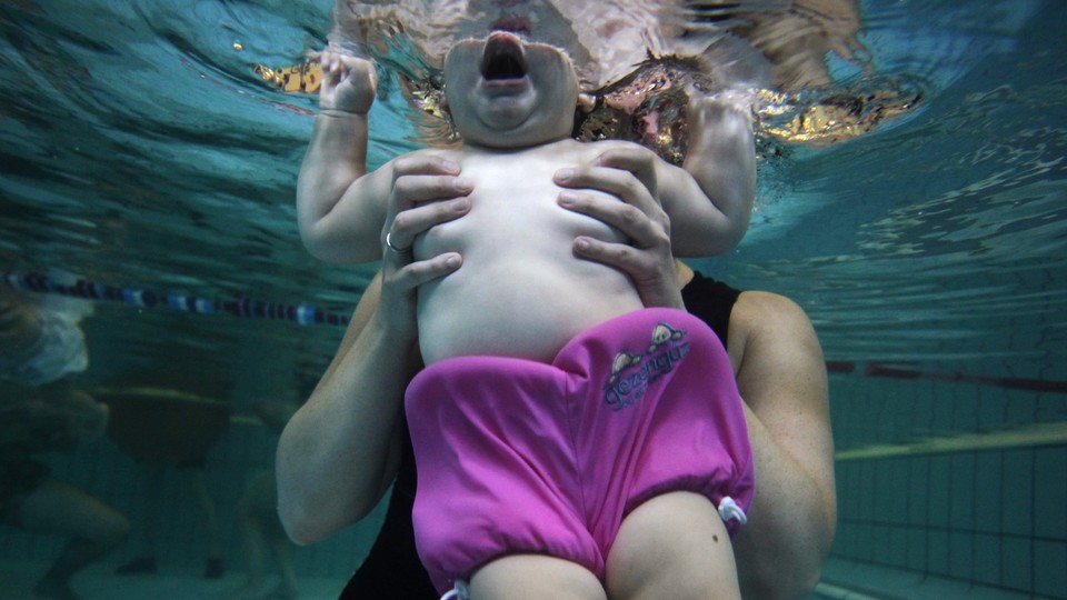 A baby is seen underwater from the chin down, held in the hands of an adult.