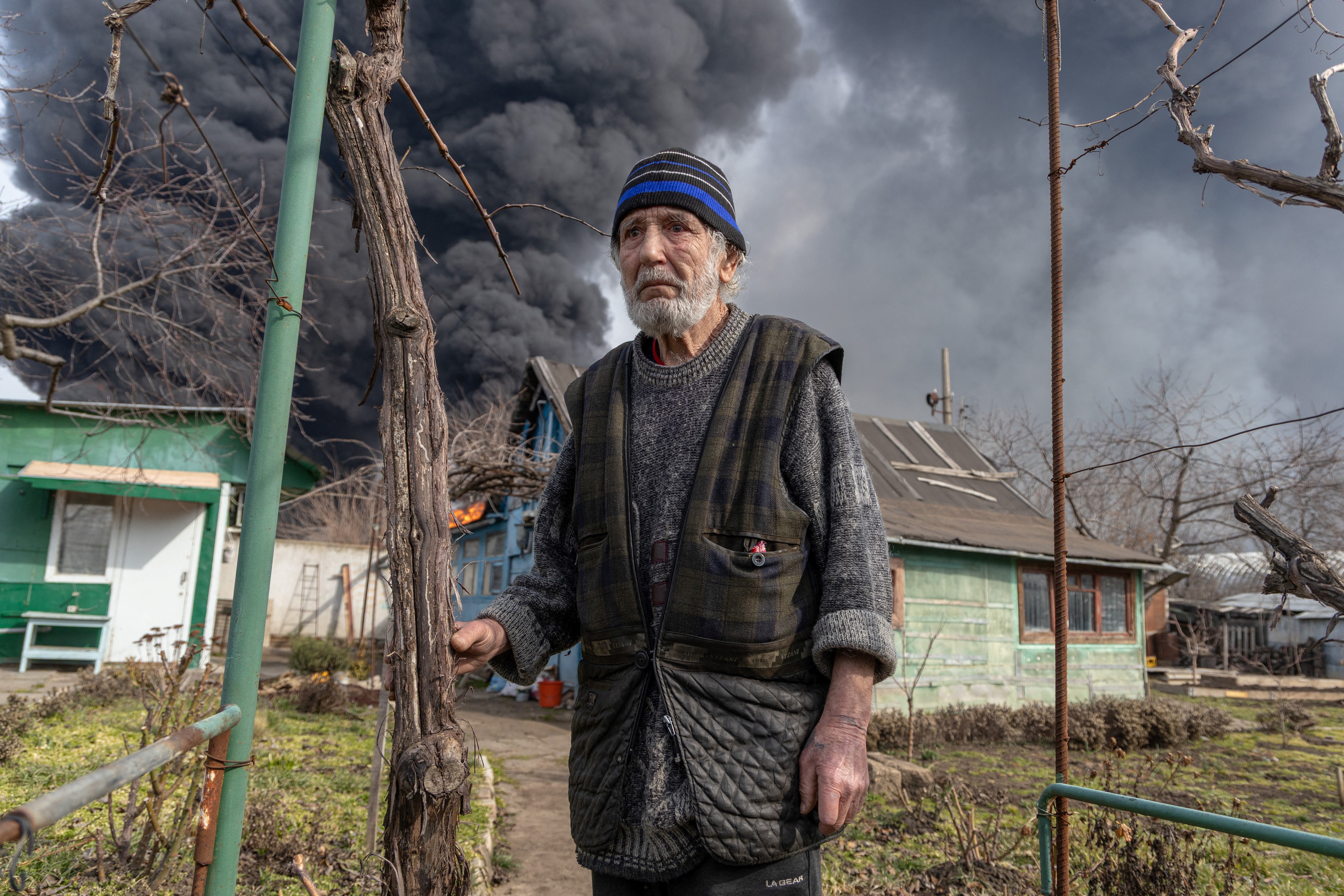 A man stands on a pathway in a courtyard outside a small house, with a large column of black smoke billowing into the air behind him.