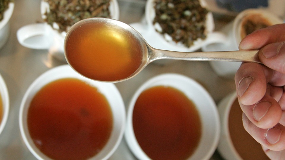 A hand holds a spoon full of tea over cups of brewed tea and tea leaves.