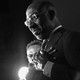 A black-and-white photo of Raphael Warnock wearing a suit and glasses in front of a spotlight, with his hands crossed over his heart