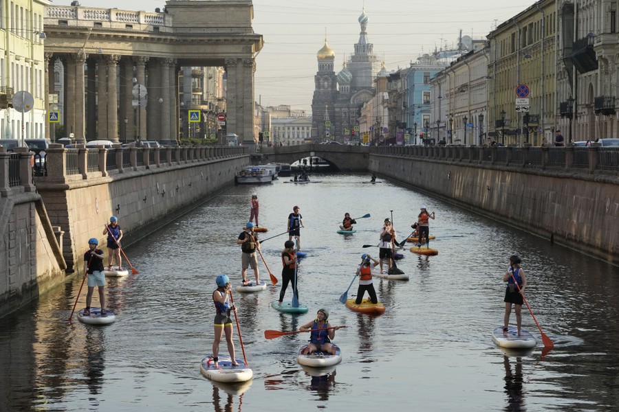 About 15 people row their paddleboards in a broad canal among the buildings of St. Petersburg.