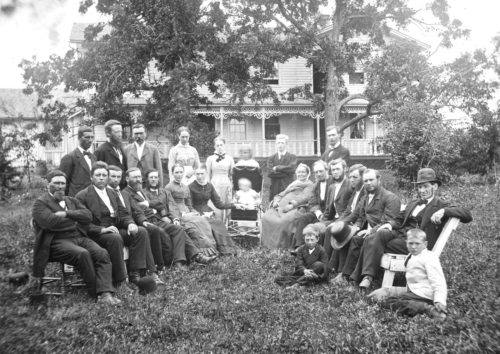 A group of twenty-four men, women, and children pose, sitting in a semi-circle in front of a large house in 1876.