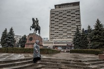 Picture of a woman wearing winter rain gear and standing up in the stairs at a square with an equestrian statue and pine trees. In the background, there is a brutalist building with a sign that reads "Cosmos."