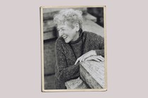 A woman with short gray hair in a gray sweater, laughing, in a photo with yellowed borders.