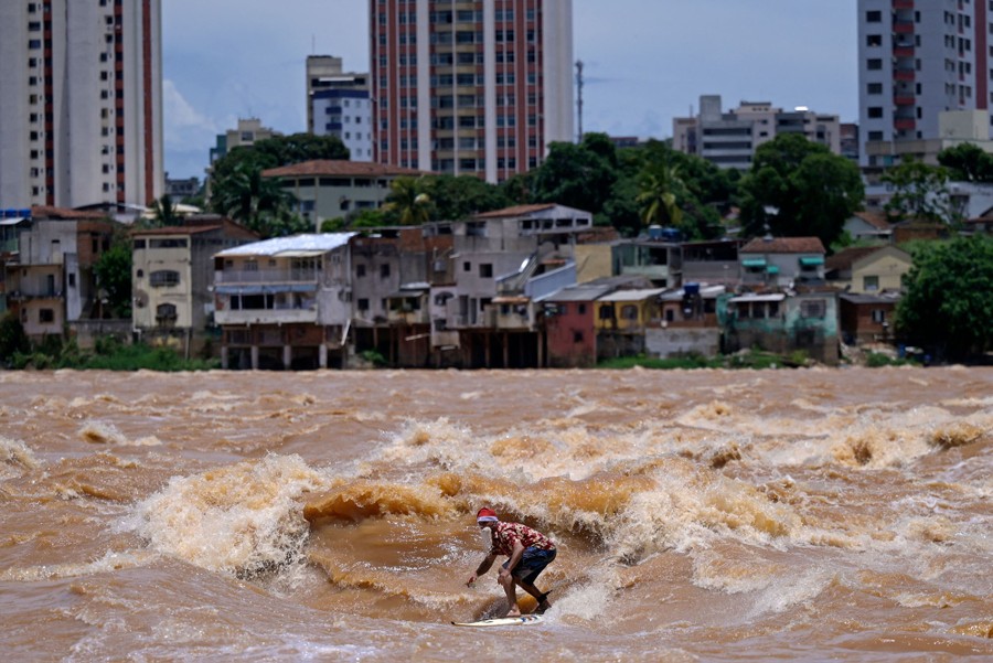 A person wearing a Santa hat and beard surfs in a choppy river, with houses and buildings in the background..