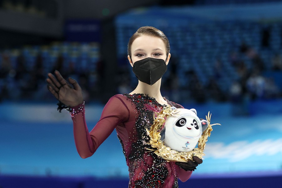 A masked figure skater waves and holds a small panda statue during a medal ceremony.