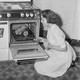 A black-and-white photo of a woman cooking a piece of meat in a gas oven