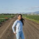 A young girl wearing a blue sweater and jeans stands on a dirt path in a field