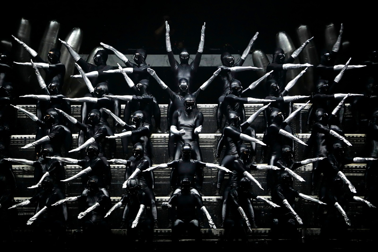 Performers on a stage, standing in tiered rows, wearing black jumpsuits with long silver gloves, pointing their arms in varying directions, creating a geometric design