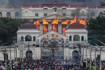 Fire and smoke rise from a large government building as protesters crowd beneath a gate.