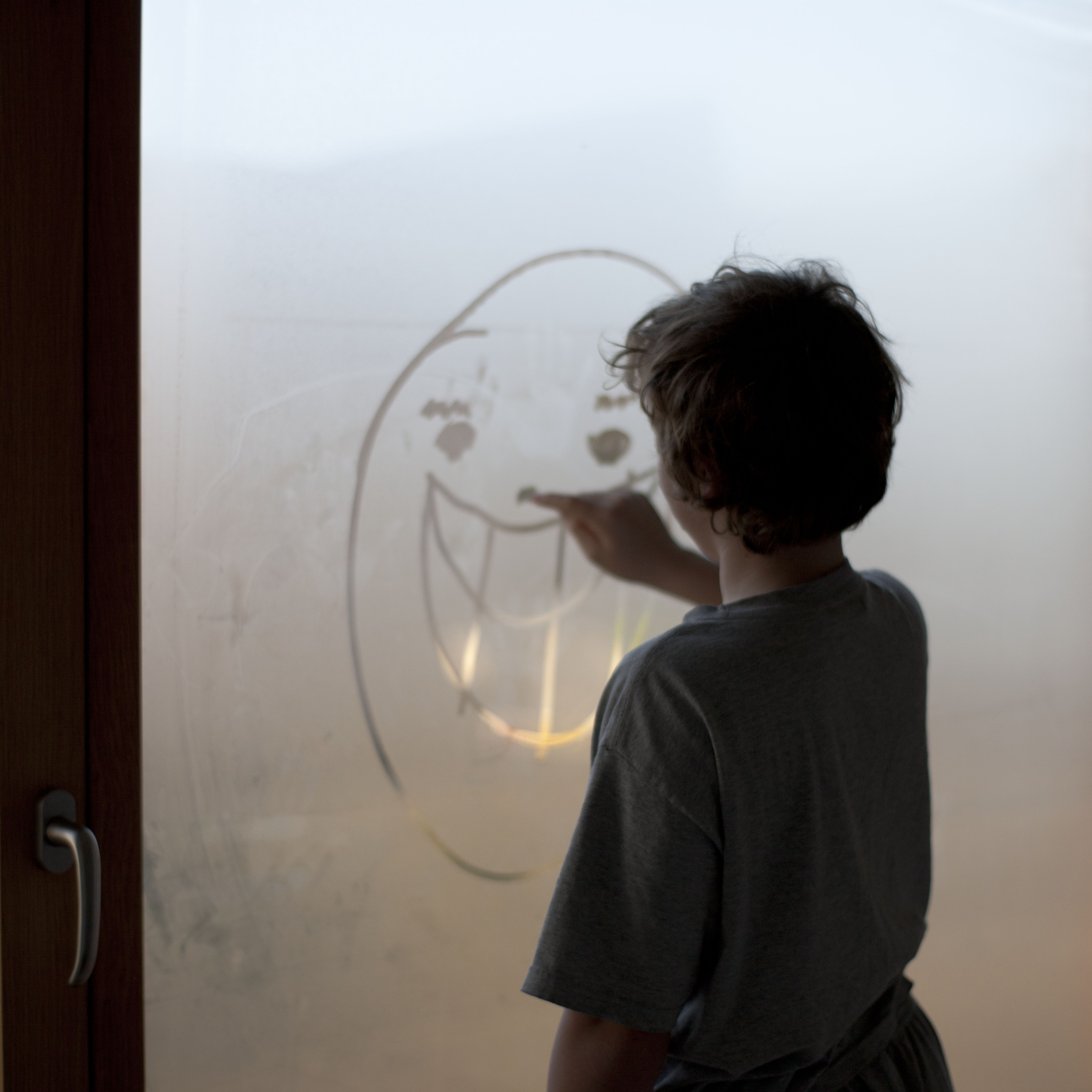 A boy drawing a face on a misted window with his finger