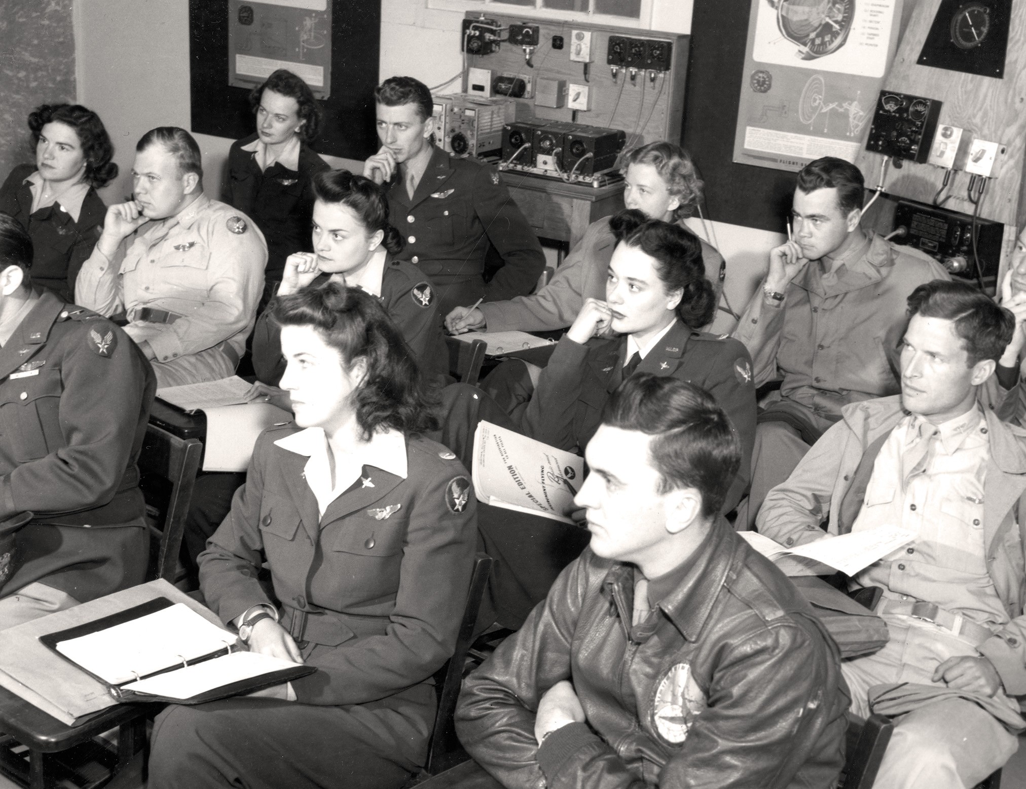 black-and-white archival photo of group of male and female pilots in uniform listening to someone speaking and sitting at classroom-style chair desks, with electronic equipment in background