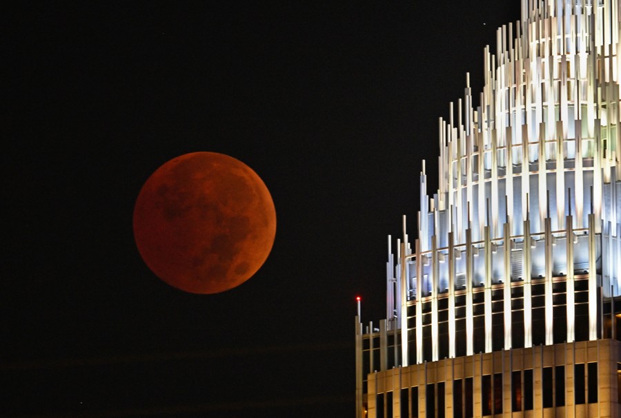 A dark-red moon beside the top of a tall building