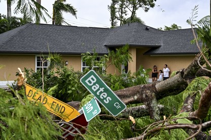 Three people stand against the wall of a house; a downed tree and street signs are in the foreground.
