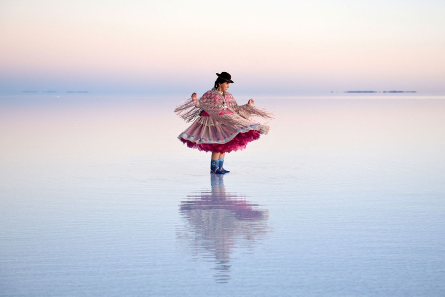 A woman wearing a black hat, an intricate, fringed shawl, and a lush, ruffled dress poses on a broad salt flat, her underskirts a pop of magenta against a pale blue-and-cream background.