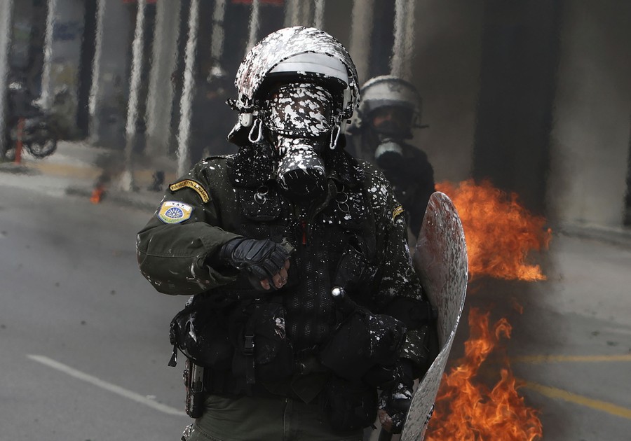 A riot policeman covered in splashed white paint walks past a flame in a street.