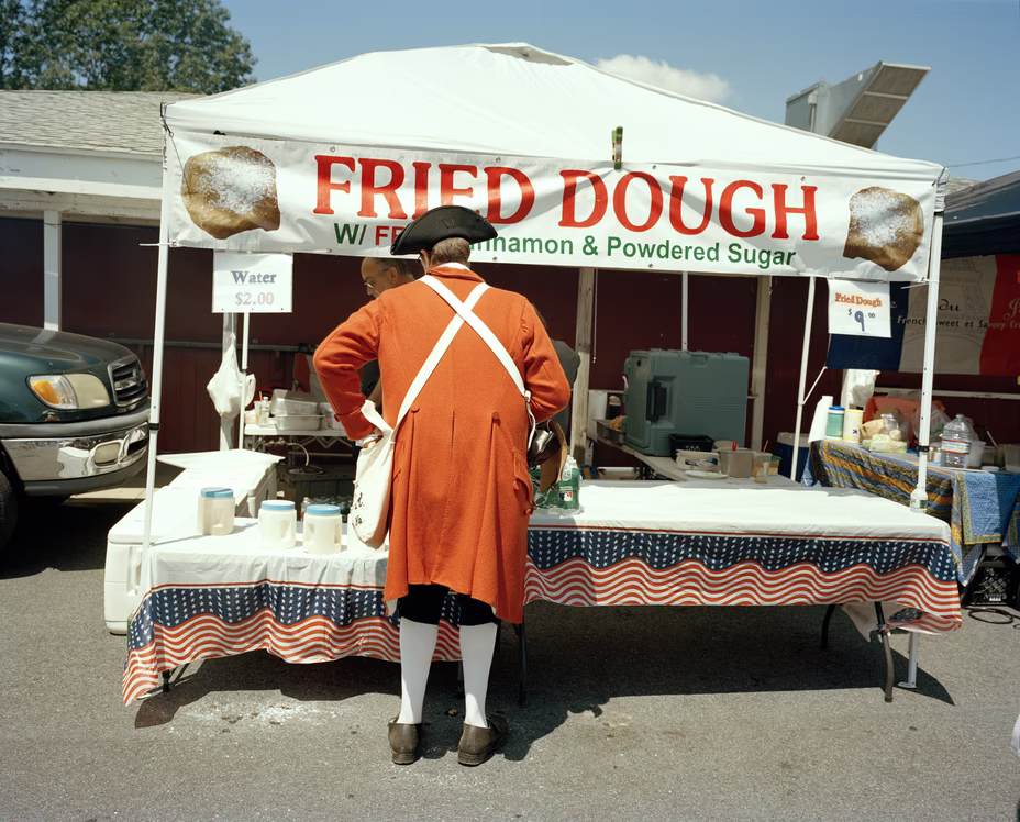 photo from behind of a reenactor playing a British soldier dressed in full regalia ordering food at a Fried Dough tent in the parking lot of the reenactment