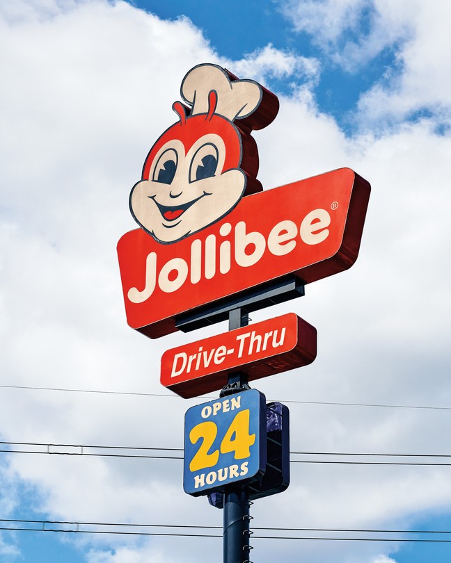 photo of roadside red Jollibee sign with smiling bee, Drive-Thru, and open 24 hours on it, with bright blue sky and clouds in background