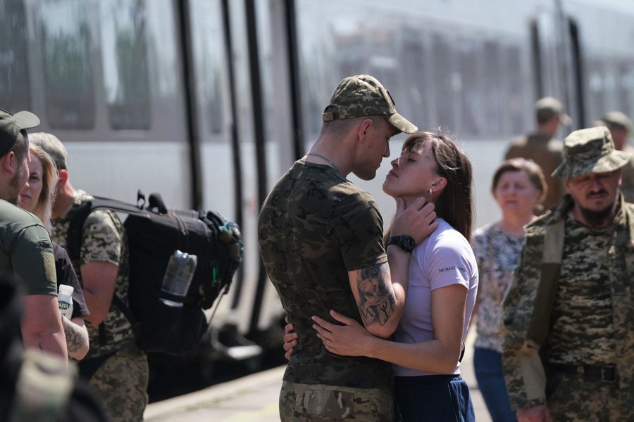 A couple embrace at a railway station, among other soldiers and civilians.