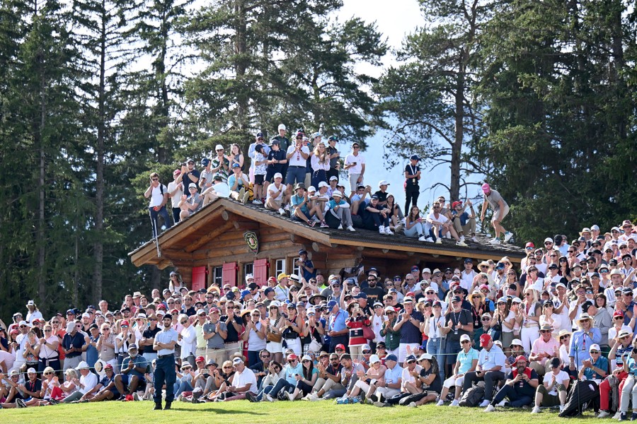 A large crowd, around and atop a small cabin, watches a golf tournament.