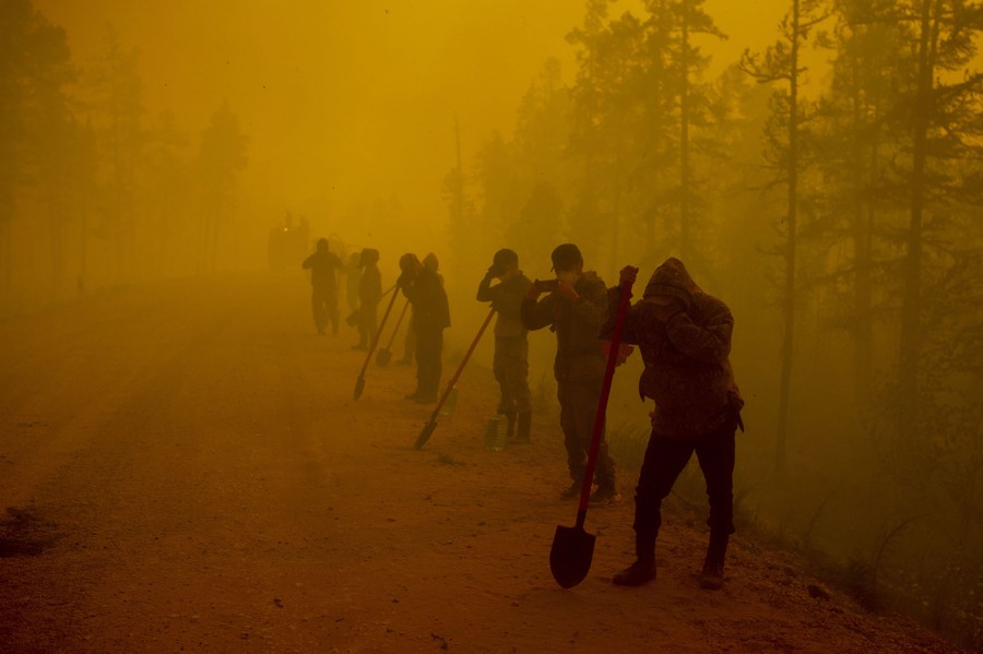 Several firefighters stand on the side of a dirt road amid thick orange smoke.