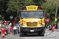 The front of a school bus fills most of the frame, as children and parents with backpacks mill about around the front of the bus. The stop sign is out on the side of the bus.